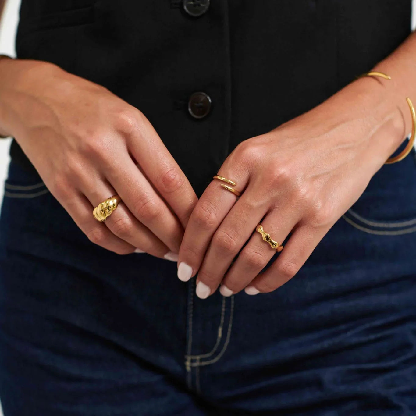 Close-up of hands wearing three gold rings and a gold bracelet, with white nail polish, black top, and blue jeans