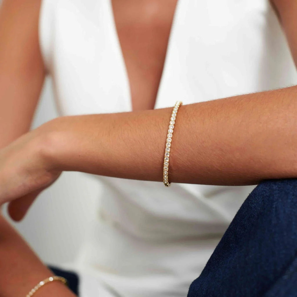 Close-up of a woman's arm wearing a gold tennis bracelet with clear gemstones, dressed in white and blue