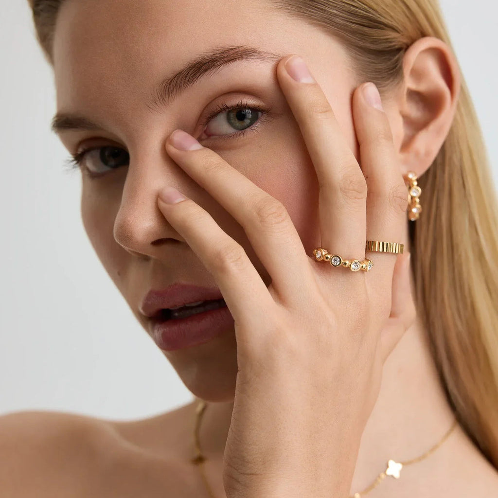 Close-up of woman wearing gold beaded tennis ring and textured band ring, with matching gold earrings