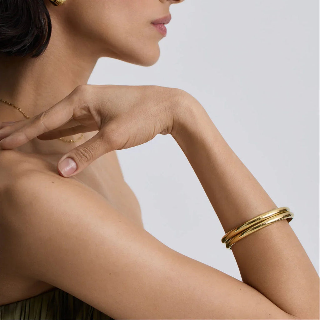 Close-up of woman wearing chunky classic gold bangles on arm with minimal jewelry and neutral background