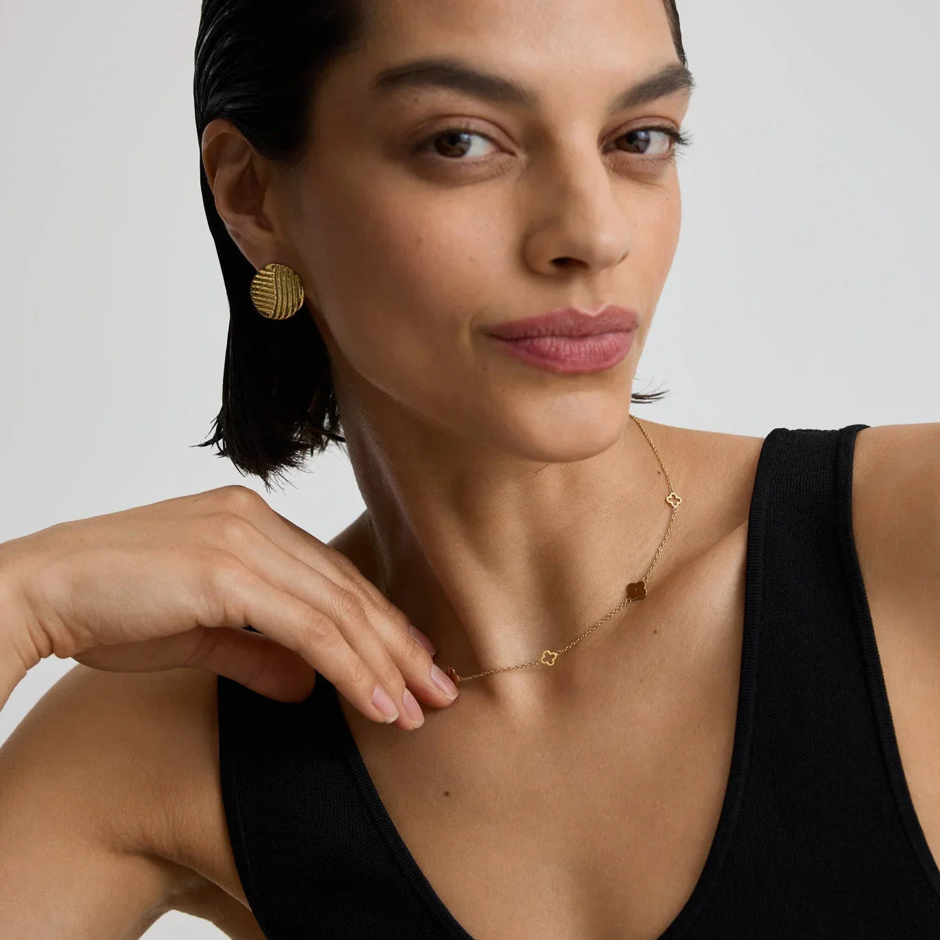 Close-up of woman wearing a delicate gold clover necklace and textured gold disc earrings with a black sleeveless top