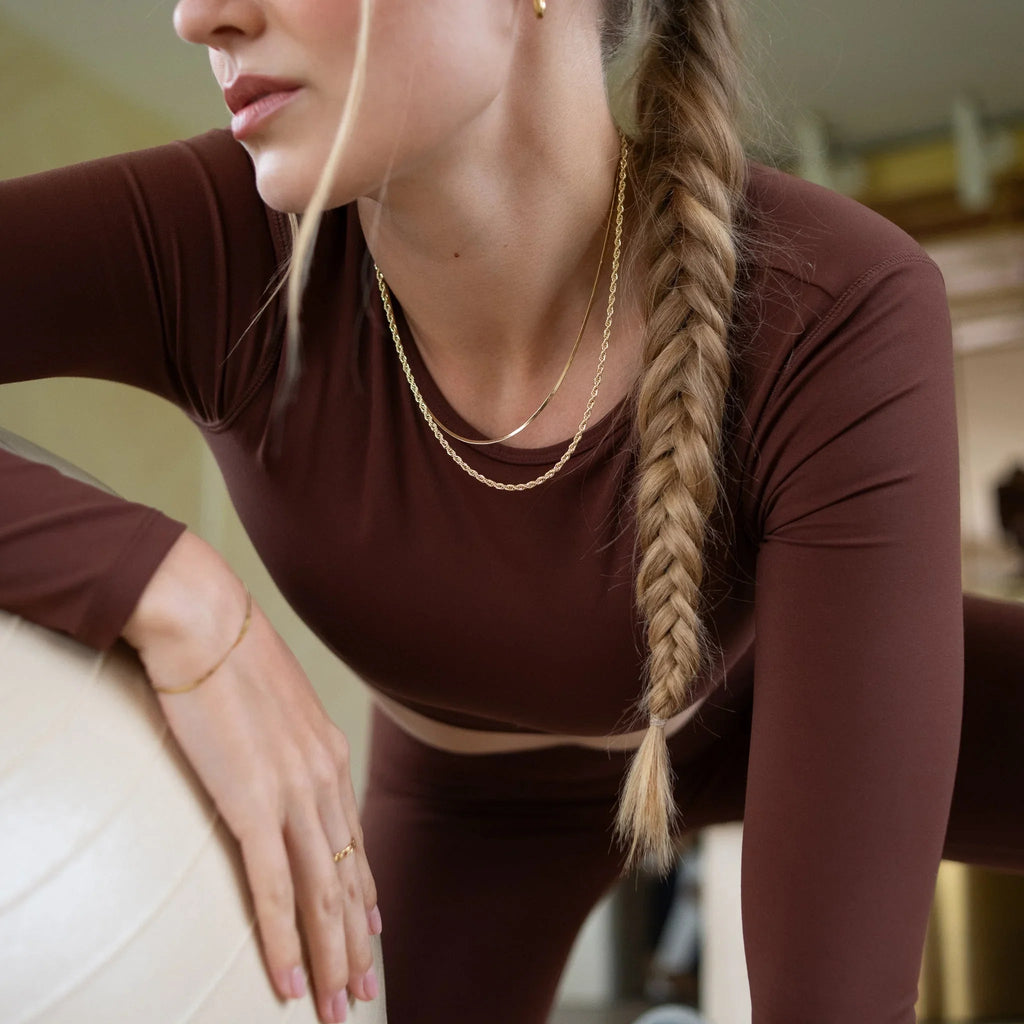 Woman with long braid wearing brown activewear and gold jewelry, leaning on a white exercise ball indoors