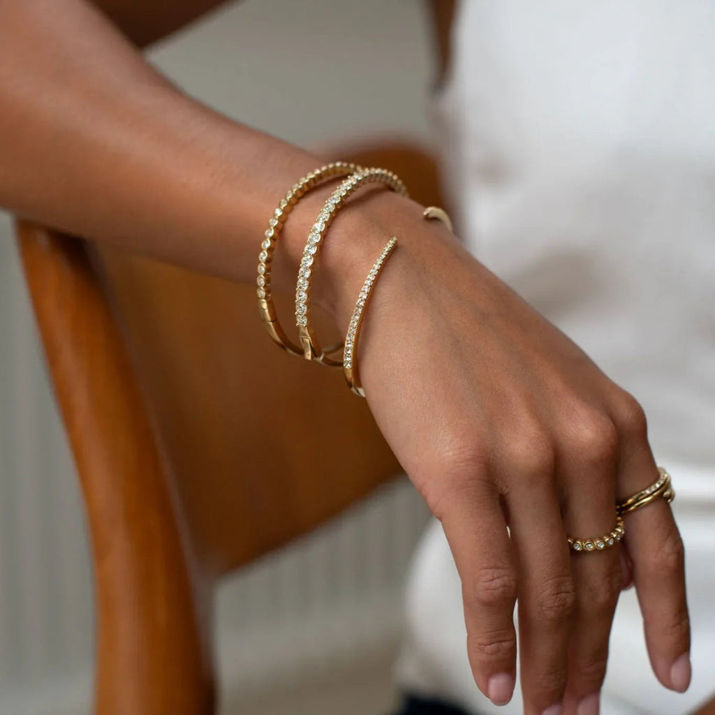 Close-up of a wrist with three gold diamond bracelets and two gold rings, hand resting on a wooden chair