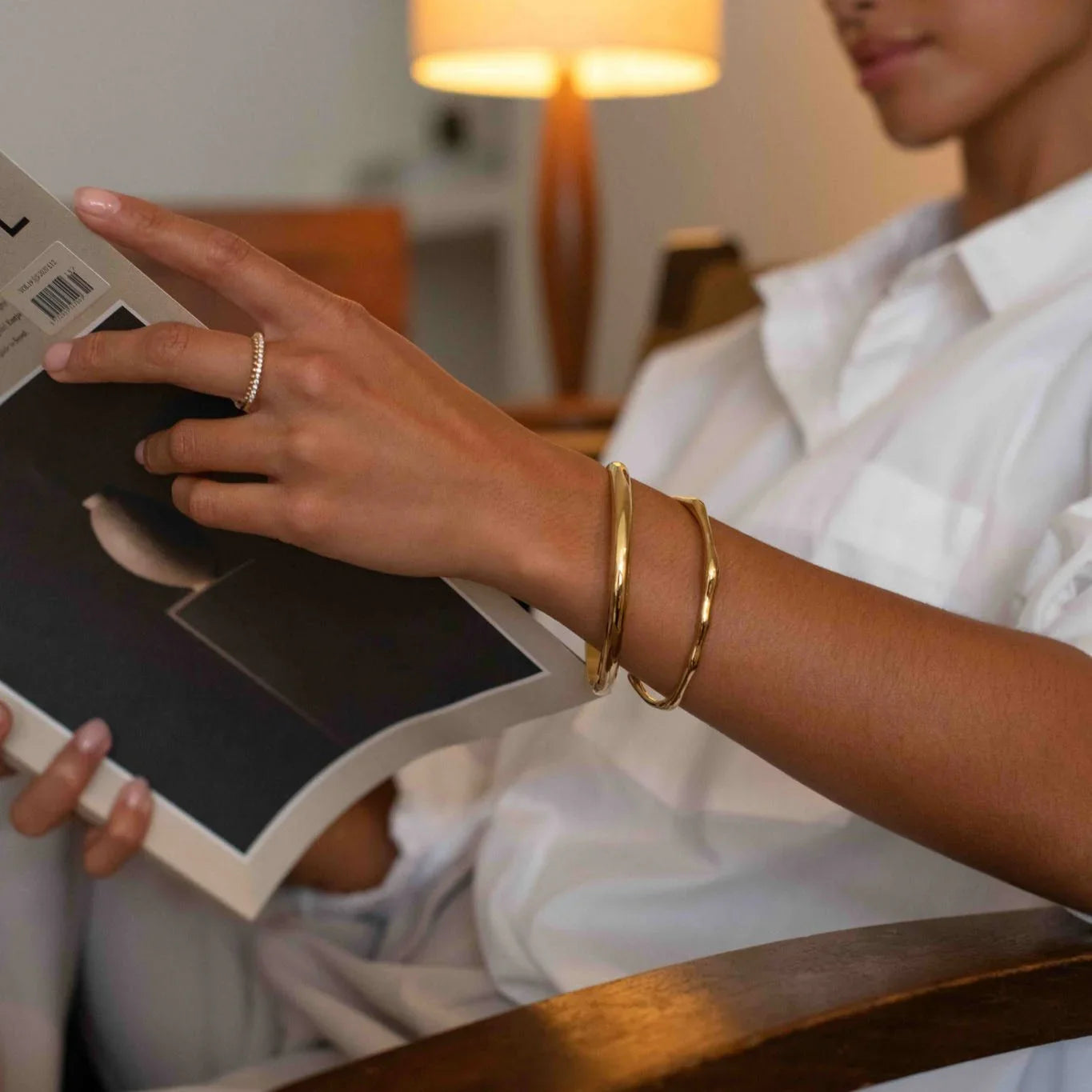 Close-up of a person in white shirt reading a book, wearing gold bracelets and a ring in cozy indoor setting