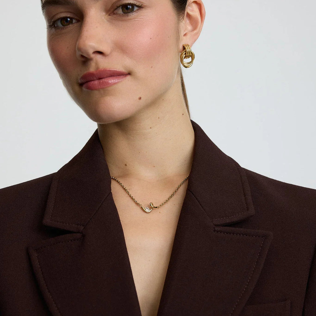 Close-up of woman wearing gold twisted hoop earrings and delicate chain necklace with curved pendant, in brown blazer