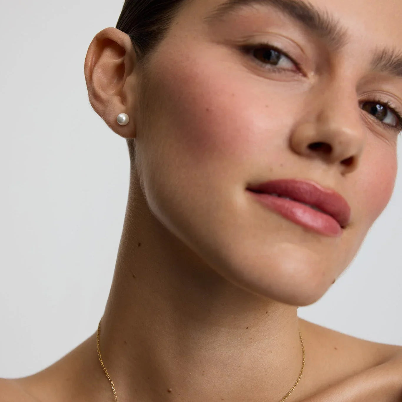 Close-up of woman wearing medium pearl stud earrings with natural makeup and gold chain necklace
